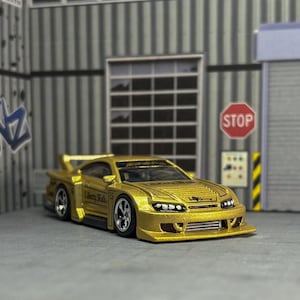 Puede incluir: Un coche de juguete dorado con alerón y ruedas personalizadas. El coche tiene el texto "Liberty Walk" y el número "15" en el lateral. El fondo es un garaje con grafitis y una señal de stop.
