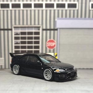 May include: A black die-cast model car featuring a rear spoiler, white multi-spoke wheels, and clear headlights. The vehicle is positioned in front of a miniature garage backdrop with a stop sign, a large closed door, and a wall with graffiti text.