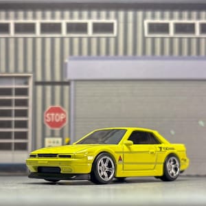 May include: A bright yellow die-cast toy car featuring a sleek coupe body style, chrome-finished multi-spoke wheels, a black front air dam, and a small Yokohama logo printed on the rear quarter panel, presented in a detailed scale model format.