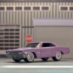 Könnte beinhalten: Ein hellviolettes Spielzeugauto aus Metall mit zweitüriger Coupé-Karosserie, silbernem Kühlergrill und weißen Rädern. Das Fahrzeug hat eine tiefe Straßenlage und ist vor einem Miniatur-Garagengebäude mit einem Stoppschild positioniert.