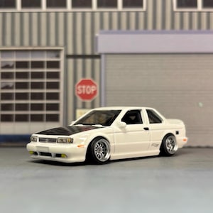 May include: A white two-door die-cast model car featuring a black hood, lowered suspension, and silver multi-spoke wheels. The vehicle has clear headlights, a front grille, and side mirrors, presented in a scale replica format with detailed body lines.