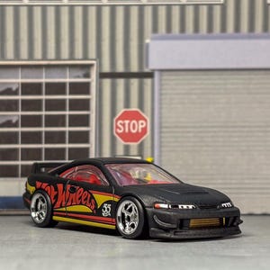 May include: A black toy car with red and yellow detailing, displaying the "Hot Wheels" logo and the number "35". The car is positioned in front of a garage-style backdrop with a stop sign.