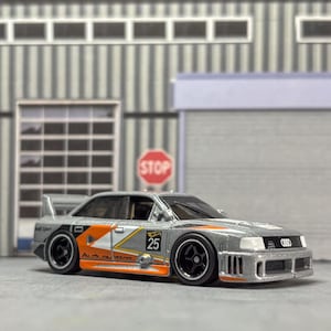 May include: A silver Audi race car toy with orange and black detailing, a rear spoiler, and black wheels. The number 25 is visible on the side. The car is set against a backdrop of a garage and a stop sign.