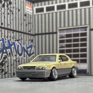 May include: A miniature beige and gray sedan car model. The car is parked in a diorama setting with graffiti art and a stop sign. The text "MCREATIONZ" is visible on the wall.