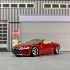 Puede incluir: Un coche de juguete descapotable rojo con interior beige. El coche tiene ruedas plateadas y está aparcado frente a un fondo industrial gris. Un letrero de stop es visible en el fondo.