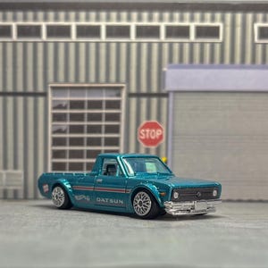 Könnte beinhalten: Ein türkisfarbenes Datsun-Pickup-Modellauto mit silbernen Rädern, einer verchromten vorderen Stoßstange und einem seitlichen Streifengrafik-Design mit dem Schriftzug Datsun. Das Fahrzeug hat einen schwarzen Kühlergrill, klare Fenster und eine Ladefläche.