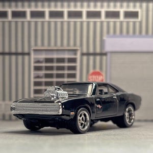 May include: A black die-cast toy muscle car featuring a prominent chrome engine protruding from the hood, a silver front grille, and chrome-rimmed wheels. The vehicle displays a red R/T emblem on the side panel and has a sleek, glossy finish.
