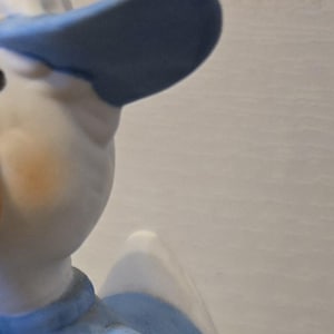 May include: A ceramic figurine of a duck wearing a blue baseball cap and jersey, holding a baseball bat and a baseball. The base of the figurine is green and has the text "Here a hit with me!" written on it.