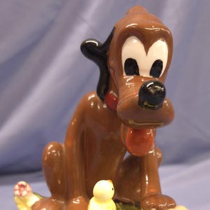 May include: A ceramic figurine of a cartoon dog with a black ear, brown body, and a red collar. The dog is sitting on a green base with a bone and a small yellow chick. The base has pink and white flowers.