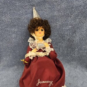 May include: A vintage doll wearing a burgundy dress with white lace trim and the word "January" embroidered on the front. The doll has brown curly hair, a silver hat, and is holding a silver object. Includes a stand and tags.