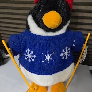 May include: A plush penguin wearing a blue sweater with white snowflakes and a red knit hat, holding yellow skis.