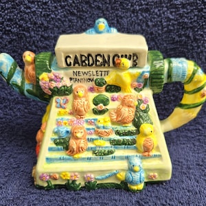 May include: A ceramic teapot with a garden theme. The teapot is decorated with owls, flowers, and the words "GARDEN CLUB NEWSLETTE PLANT NOW". The handle and spout are yellow and green, and the base is light blue.
