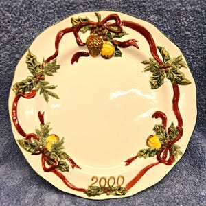 May include: Cream-colored decorative plate with a scalloped edge. The plate features a red ribbon design with holly leaves, berries, and pinecones. The year "2000" is displayed at the bottom of the plate.