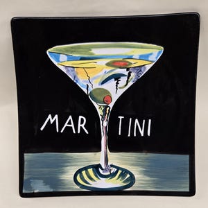 May include: Square black ceramic plate with a painted martini glass design. The glass contains olives and a red garnish. The word "MARTINI" is written in white. The base of the glass sits on a blue and green painted surface.