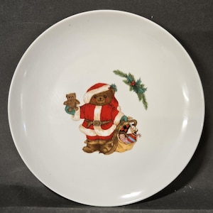 May include: A white ceramic plate featuring a central illustration of a brown teddy bear dressed in a red and white Santa Claus suit. The bear holds a small teddy bear and carries a sack filled with toys, with a sprig of green holly above it.