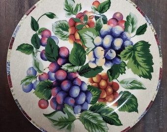 May include: A white ceramic dinner plate with a decorative design of grapes and leaves. The grapes are purple, red, and green, and the leaves are green. The plate has a brown, red, and white border.