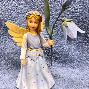May include: A figurine of a young angel with blonde hair, yellow wings, and a light blue dress. The angel holds a blue flower with a green stem. The angel has a floral crown and is standing on a blue surface.