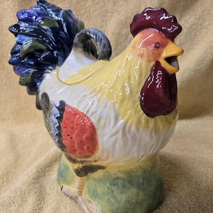 May include: A ceramic rooster teapot with a yellow body, red comb, and blue and green tail feathers. The teapot has a lid and a spout. The rooster is standing on a green base.
