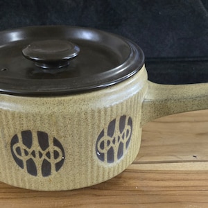 May include: A tan ceramic saucepan with a dark brown lid and handle. The pot features a repeating pattern of brown abstract designs. The handle is integrated into the pot's design, and the lid has a small knob.