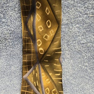 May include: A patterned necktie with a geometric design. The tie features a repeating pattern of squares, diamonds, and rectangles in shades of brown, gold, and gray. The tie is made of a smooth, possibly silk, material.
