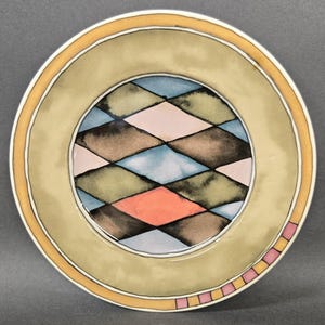 May include: A round ceramic plate featuring a central pattern of interlocking diamonds in shades of blue, pink, brown, and green. The wide rim is a muted olive green with a thin yellow border and a small section of pink and yellow squares.