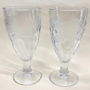 May include: Two clear, stemmed glasses with etched floral designs. The glasses have a tulip shape, with a wide bowl that tapers to a narrow stem and a round base. The etched design features flowers and leaves. The glasses are made of clear glass.