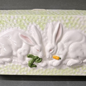 May include: A rectangular ceramic box with a light green textured background. Two white rabbits with pink ears are in the center, facing each other. One rabbit holds a green leaf, the other a small orange carrot. The box has a white lid.