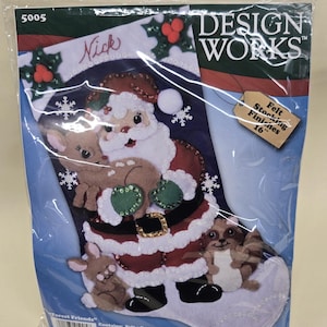 May include: A Christmas stocking kit featuring a felt design with Santa Claus, a reindeer, a rabbit, and a raccoon. The stocking is white with red and green accents, and the text "Nick" is visible. The kit is labeled "Design Works" and is 16 inches in length.