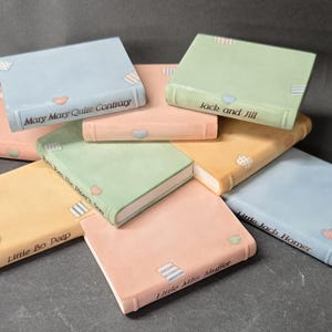 May include: A collection of eight rectangular ceramic book-shaped ornaments in pastel shades of blue, green, peach, and yellow. Each piece features a nursery rhyme title printed on the spine, including Mary Mary Quite Contrary, Jack and Jill, and Little Bo Peep.