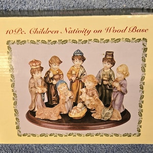 May include: A boxed set of a 10-piece children's nativity scene on a wood base. The figures are dressed in various robes and garments, with a baby in a manger. The box has the text "10 Pc. Children Nativity on Wood Base".