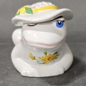 May include: A white ceramic frog figurine wearing a wide-brimmed hat adorned with yellow flowers and a yellow ribbon. The frog has one blue eye open and one eye closed in a wink, with a painted yellow floral design on its chest and a pink mouth.