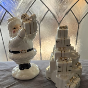 May include: White ceramic salt and pepper shakers in the shape of a Santa Claus holding a gift and a stack of presents. The Santa Claus shaker has a black belt and a black hat. The presents shaker has gold accents.