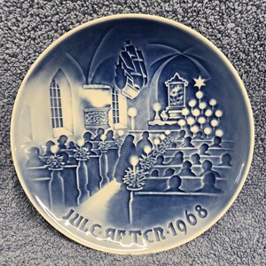 May include: A blue and white decorative plate featuring a church scene with figures, a ship, and a Christmas tree. The plate has the text "JULE AFTEN 1968" around the bottom edge. The plate is round and has a raised relief design.