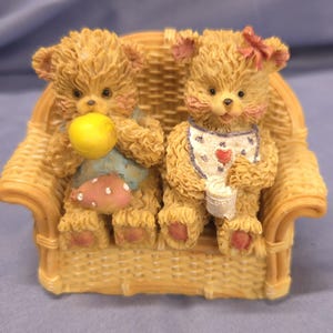 May include: A figurine of two teddy bears seated on a wicker sofa. One bear holds a yellow ball and a pink mushroom, the other wears a bib and holds a cup. The bears have pink accents and are light brown in colour.