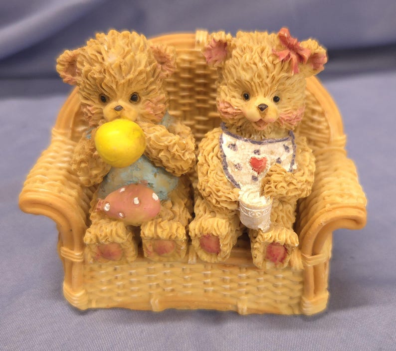 May include: A figurine of two teddy bears seated on a wicker sofa. One bear holds a yellow ball and a pink mushroom, the other wears a bib and holds a cup. The bears have pink accents and are light brown in colour.