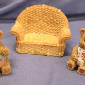 May include: A miniature wicker sofa with two teddy bear figurines. One bear holds a yellow ball and wears a bib with a pink polka-dot design. The other bear wears a bib with a heart and a pink bow.