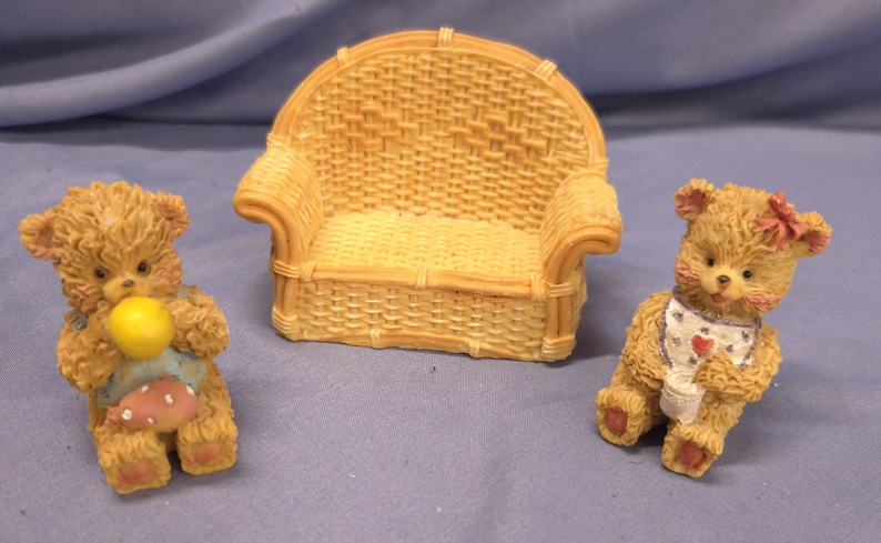 May include: A miniature wicker sofa with two teddy bear figurines. One bear holds a yellow ball and wears a bib with a pink polka-dot design. The other bear wears a bib with a heart and a pink bow.
