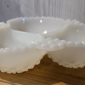 May include: A white, three-sectioned serving dish with a scalloped edge and a textured, hobnail-like pattern. The dish is made of opaque glass and sits on a wooden surface.