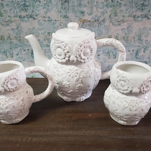 May include: A white ceramic owl-shaped tea set, including a teapot with a lid and two mugs. The set features floral and owl face detailing. The background is a patterned blue and white design.