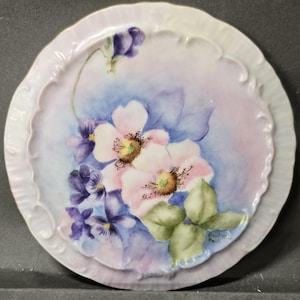 May include: A round porcelain decorative plate with a scalloped, embossed edge. The center features a hand-painted floral design of two large pale pink flowers with yellow centers, surrounded by clusters of small purple violets and green leaves.