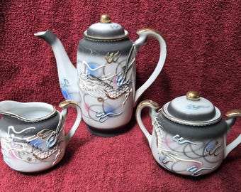 May include: A three-piece porcelain tea set with a black and white design, featuring a teapot, creamer, and sugar bowl. The set is decorated with a dragon motif, gold accents, and colorful swirls.