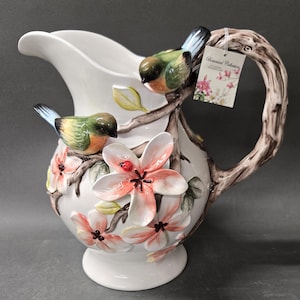 May include: A white ceramic pitcher featuring three-dimensional sculpted details of two small birds with green and blue wings perched on brown branches. The pitcher is decorated with pink flowers and a small red ladybug, and includes a hanging tag.