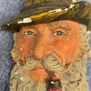 May include: A detailed, hand-painted figurine of an elderly man with a long, white beard and mustache, wearing a green hat. He is holding a pipe with red cherries attached. The figurine has a weathered appearance.