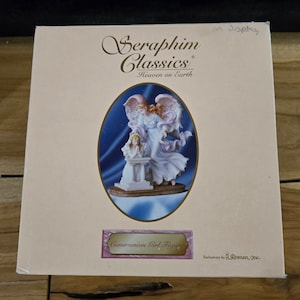 May include: A white porcelain figurine of an angel with wings outstretched, holding a small girl in a white dress. The girl is kneeling and praying. The figurine is on a blue and white base with the words "Seraphim Classics Heaven on Earth" and "Communion Girl Figure" printed on the box.