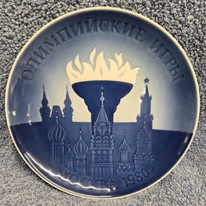 May include: A blue and white decorative plate featuring a torch with flames above a skyline of buildings. The plate has the text "ОЛИМПИЙСКИЕ ИГРЫ" and "МОСКВА 1980" around the rim. The plate is round.