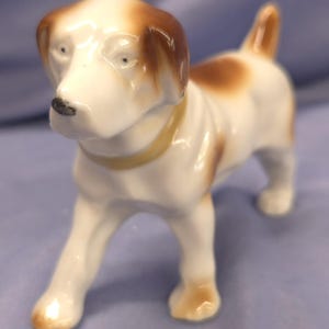 May include: A ceramic dog figurine in a standing pose. The dog has a white body with brown patches on its head and back. It has a yellow collar and a black nose. The figurine is detailed and appears to be vintage.