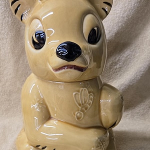 May include: A ceramic bear cookie jar with a light brown glaze and black accents. The bear is sitting with its paws crossed and has a sweet expression on its face. The jar is decorated with small bee designs.