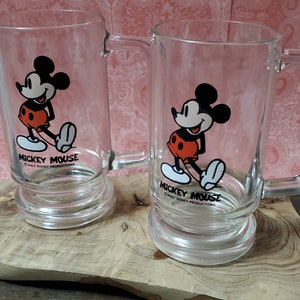 May include: Two clear glass mugs with Mickey Mouse designs. Each mug features a red and black image of Mickey Mouse with the text "Mickey Mouse" and "© Walt Disney Productions" printed on the front.