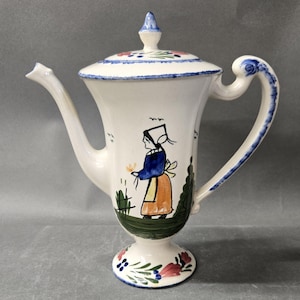 May include: A cream-colored ceramic teapot featuring a hand-painted illustration of a person in a blue top and orange skirt standing in a green field. The teapot has a matching lid, a curved handle, a long spout, and floral accents in blue and pink.