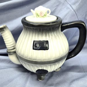 May include: A ceramic teapot with a white and gray color scheme. The teapot has a black handle and spout, and a small teapot-shaped lid with a pink rose design. The body of the teapot has vertical ridges.